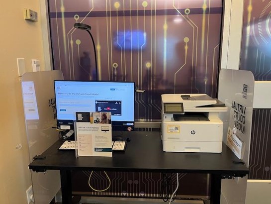 black desk with a computer monitor, a thin keyboard, a printer/scanner, and a plastic display holder for 8x10 paper. The content showing on the monitor and on the paper in the plastic display features information on virtual court services