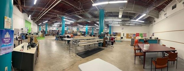 wide open space room; columns; large quilting table; laser cutter and leather making equipment/ shelf with board games; two rectangular tables pushed together with chairs; back wall shows bulleting boards with informational flyers.
