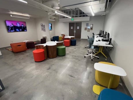 large open space room with gray cement-like floor; colorful soft seating ottomans and geometrical tables; computers on tables that are aligned against the right wall; rolling chairs; two television nook areas with rugs and lounge seating to the left; TVs are showing KPop videos;
