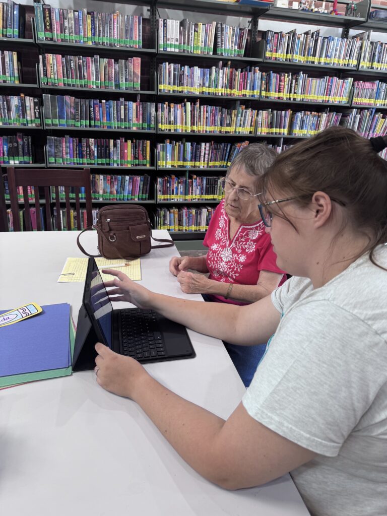A digital navigator sits at a table with a senior citizen in Martindale Community Library, offering instruction on a laptop.
