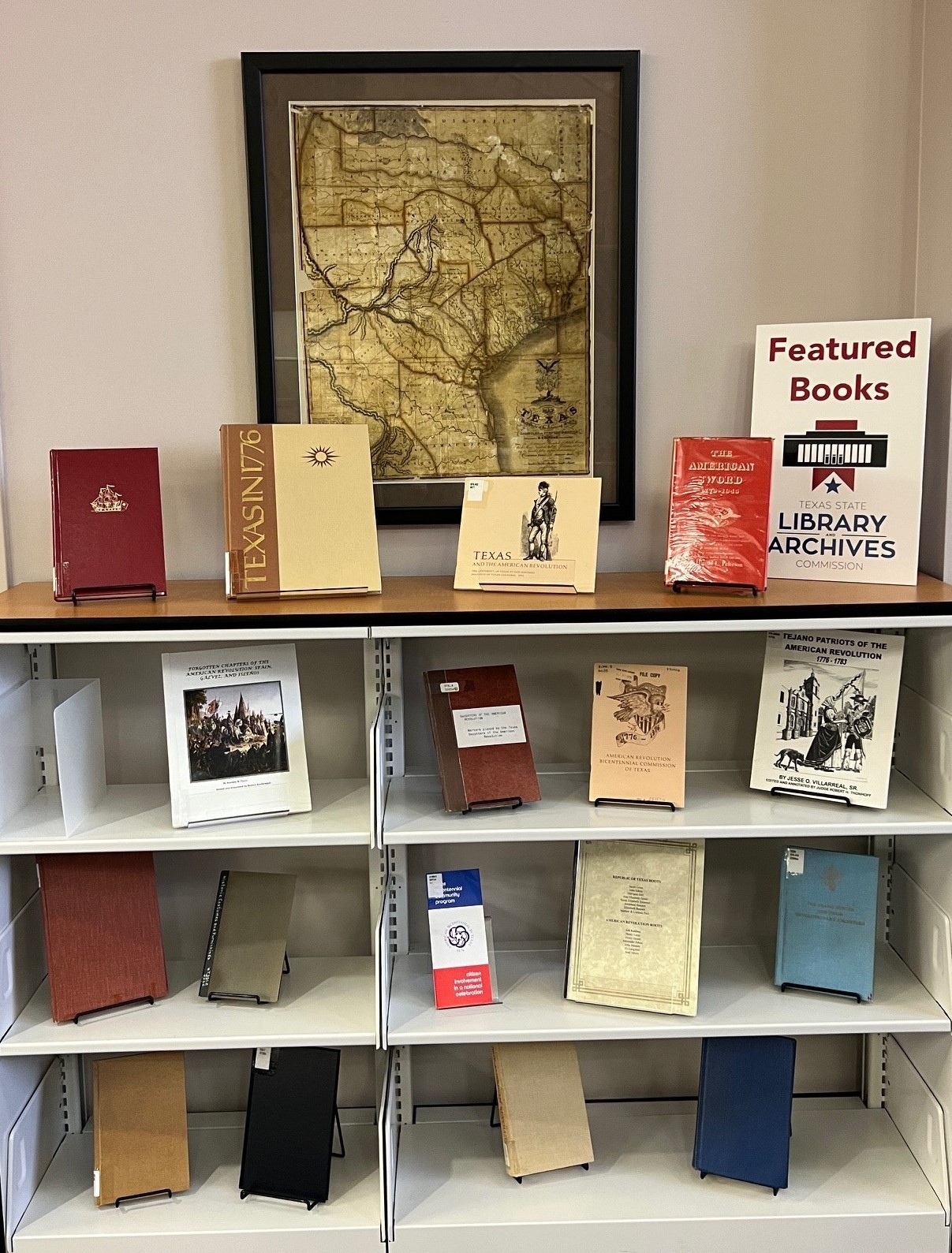 Photo of bookcase with three shelves of books facing outward and four books on top with a Featured Books sign. An antique map of Texas is hung on the wall behind the display.
