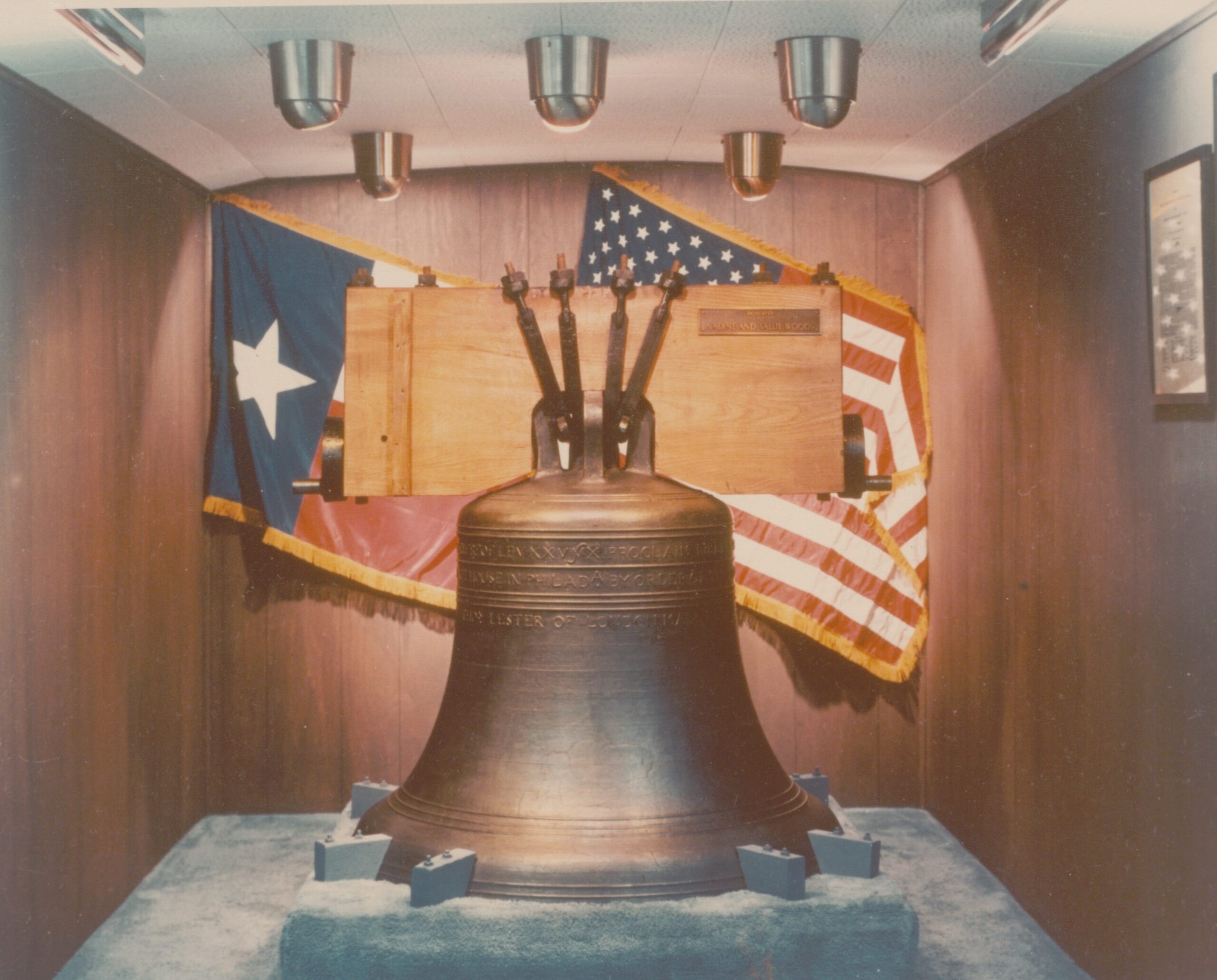 Colorized photo of Liberty Bell replica with Texas and American flags hanging behind. The bell sits on a mount and a closed space, which was in a truck called the Liberty Liner.