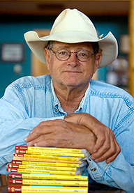 Photograph of John Erickson wearing a cowboy hat and hands crossed on a stack of books Photograph of John Erickson wearing a cowboy hat and hands crossed on a stack of books